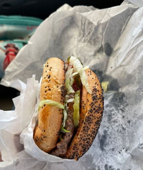 A close-up of a poppy seed bun hamburger with lettuce, ketchup, and a beef patty, wrapped in white paper.