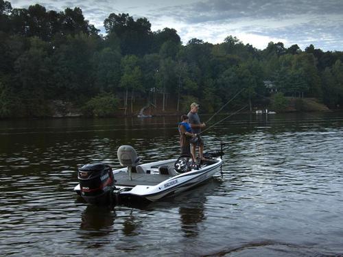 A man and a boy fishing from a small boat on a calm lake surrounded by trees under a cloudy sky.