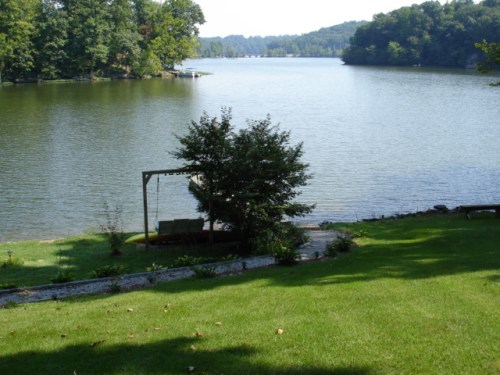 A serene lake view surrounded by trees, with a grassy area and a swing set near the water's edge.