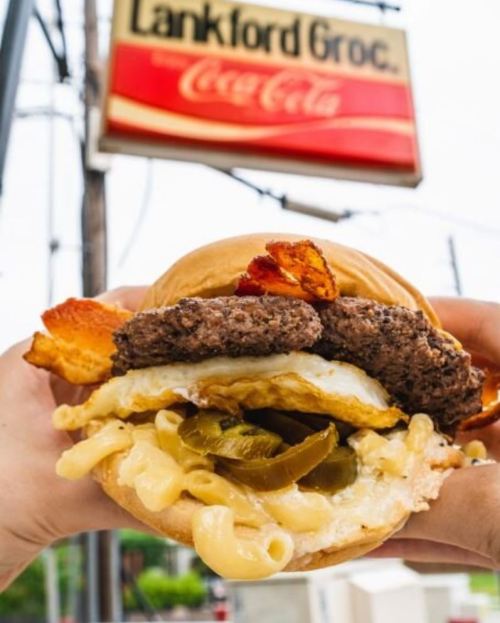 A close-up of a hearty burger with bacon, fried egg, macaroni, and pickles, held in front of a Lankford Grocery sign.