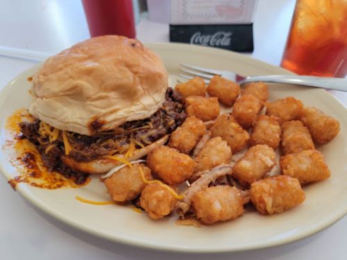 A plate with a pulled pork sandwich topped with cheese and a side of crispy tater tots.