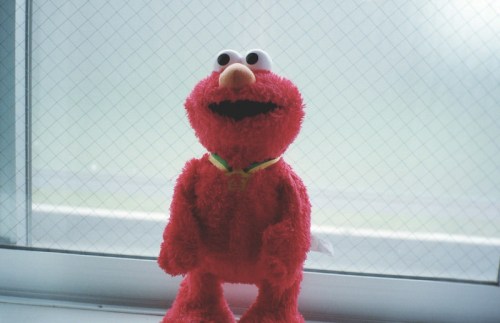 A plush toy of a red character with large eyes and a wide smile, standing by a window with a blurred background.