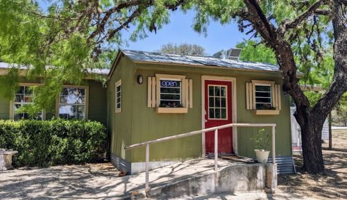 A small green building with a red door and "open" sign, surrounded by trees and a gravel path.