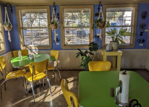 Bright, cozy café interior with green tables, yellow chairs, and large windows showcasing plants and natural light.