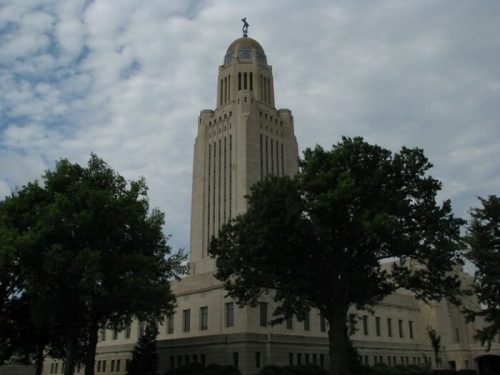 Nebraska State Capitol building with a golden dome, surrounded by trees under a cloudy sky.