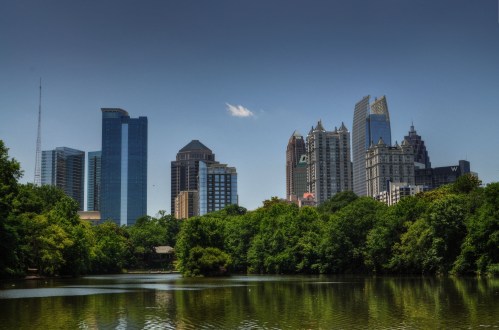City skyline with modern skyscrapers reflected in a serene lake, surrounded by lush green trees under a clear blue sky.