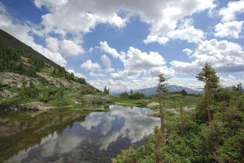 A serene landscape featuring a calm lake reflecting clouds and mountains, surrounded by lush greenery and trees.