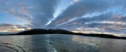A panoramic view of a calm lake at sunset, with dramatic clouds reflecting in the water.