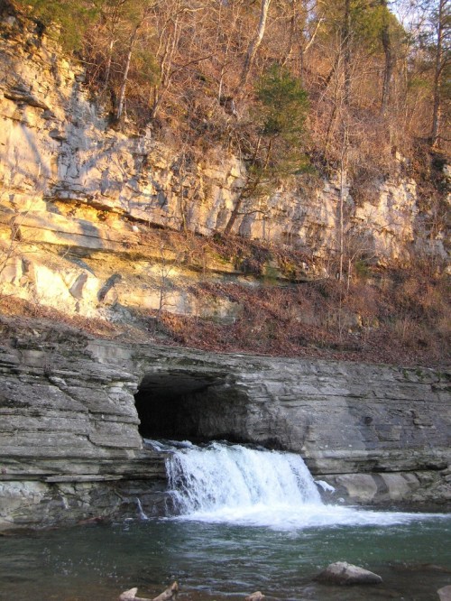A small waterfall cascades over rocky terrain, surrounded by steep cliffs and trees in a serene natural setting.