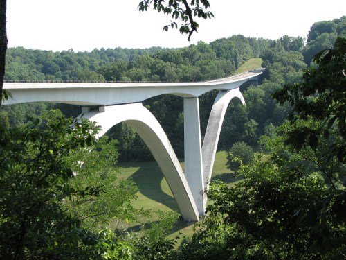 A modern concrete bridge with arch supports spans a lush green valley, surrounded by trees and hills.