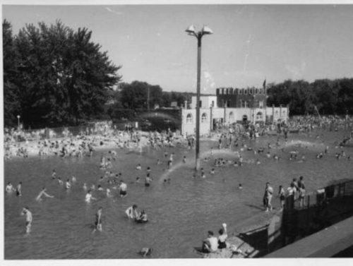 A crowded beach scene with people swimming and playing in the water, featuring a building in the background.