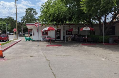A small diner with outdoor seating, red umbrellas, and a dinosaur statue in the parking lot on a sunny day.