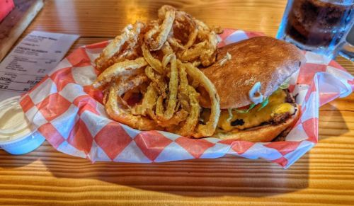 A burger with cheese and toppings served with a side of crispy onion rings in a red and white checkered basket.
