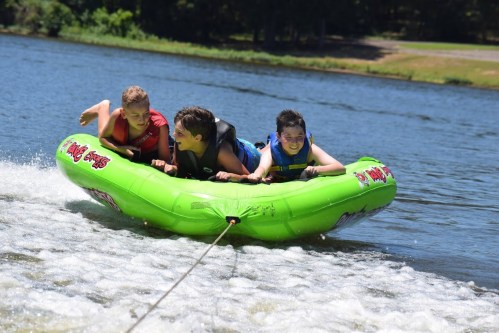 Three kids enjoy tubing on a bright green inflatable in a lake, being pulled by a boat, with splashes of water around them.