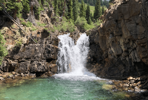 A serene waterfall cascades over rocky cliffs into a clear, turquoise pool surrounded by lush greenery.