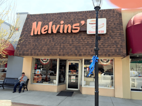 Exterior of Melvin's restaurant with a brown shingled roof, sign, and a person sitting on a bench nearby.