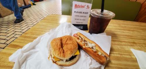 A sandwich with cheese and a hot dog topped with onions on a table, next to a drink and a "Please Wait" sign.