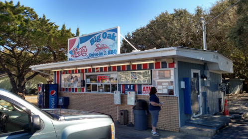 A small drive-in BBQ restaurant with a colorful sign, outdoor seating, and a customer at the counter.