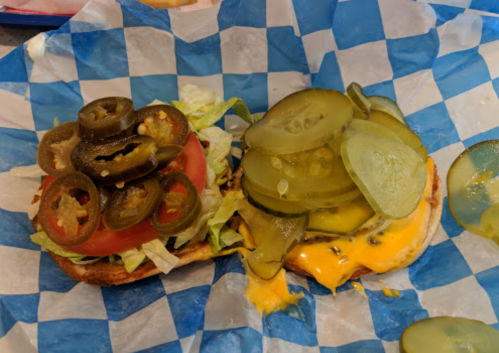 A sandwich on blue and white checkered paper, topped with lettuce, tomato, jalapeños, and pickles on a cheesy base.