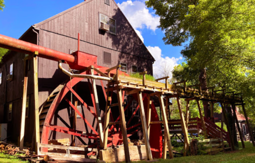 A large red waterwheel beside a wooden building, surrounded by trees and blue sky with fluffy clouds.