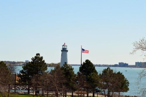 A lighthouse beside a lake, with an American flag flying and trees in the foreground against a clear blue sky.