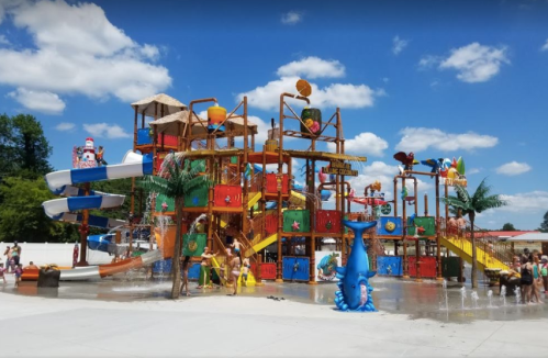 Colorful water play structure with slides, fountains, and children playing under a blue sky with fluffy clouds.