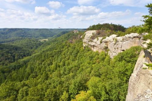 A scenic view of a rocky cliff overlooking a lush green valley and distant hills under a partly cloudy sky.