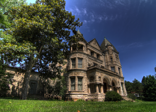 A grand, historic mansion with intricate stonework, surrounded by lush greenery and a clear blue sky.