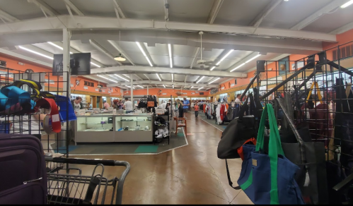 A spacious thrift store interior with clothing racks, display cases, and shoppers browsing among colorful merchandise.