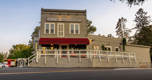 A charming gray restaurant with a red awning, surrounded by trees and a white fence, set against a clear sky.