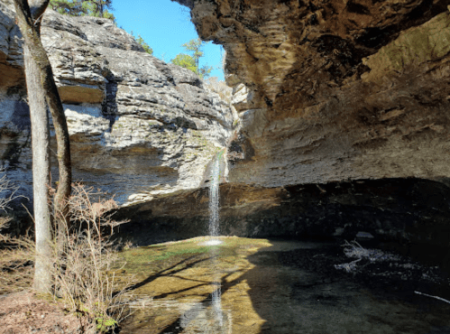 A serene waterfall cascades from a rocky overhang into a clear pool, surrounded by trees and natural rock formations.