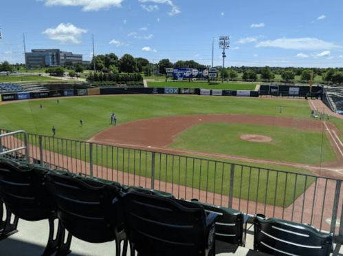A view of a baseball field from the stands, with players on the field and a clear blue sky above.