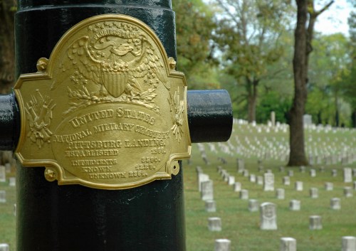 A close-up of a plaque on a cannon at a military cemetery, with rows of gravestones in the background.