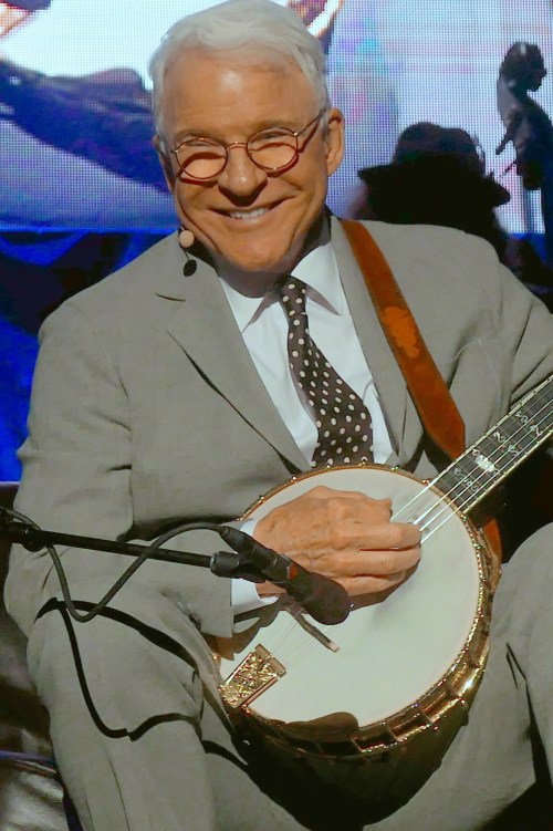A smiling man in a gray suit plays the banjo on stage, wearing round glasses and a polka dot tie.