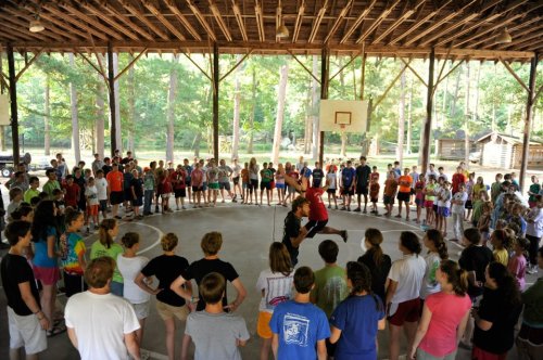 A group of children and teens watch as two performers demonstrate a basketball trick in a camp gym.