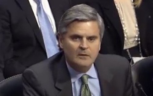 A man in a suit speaks during a formal meeting, with blurred figures in the background.