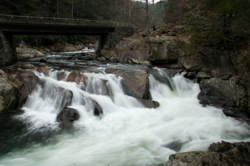 A serene waterfall cascades over rocks beneath a bridge, surrounded by lush greenery and rugged terrain.