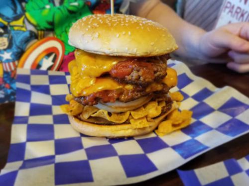 A towering burger with cheese, fried chicken, and Fritos on a blue checkered plate, with a sesame seed bun on top.