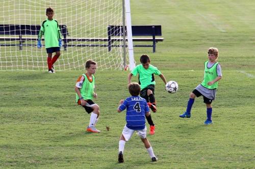 A youth soccer game with four players in green jerseys and one in blue, focused on the ball near the goal.