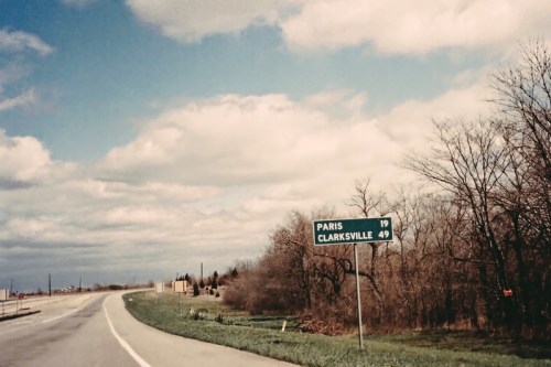 Road sign indicating distances to Paris (19 miles) and Clarksville (49 miles) with a cloudy sky in the background.