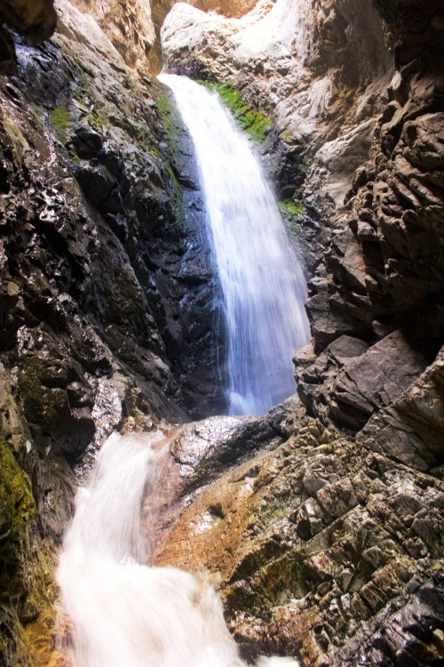A cascading waterfall flows down rocky cliffs, surrounded by textured stone and greenery. Sunlight filters through the top.