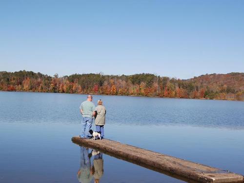 A couple and a dog stand on a dock by a calm lake, surrounded by colorful autumn trees under a clear blue sky.