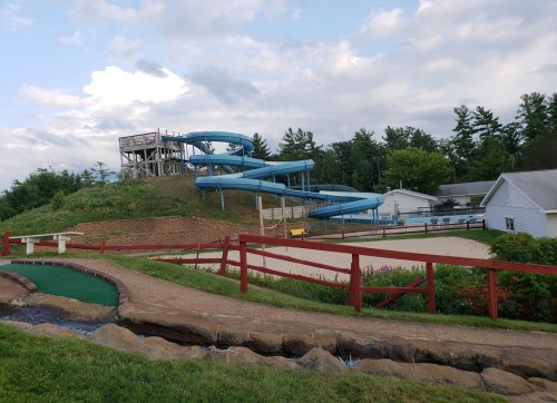 A blue water slide on a grassy hill, with a mini-golf course and sandy area nearby, under a cloudy sky.