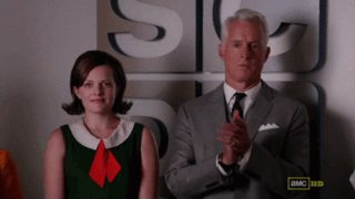 A man in a gray suit and a woman in a green dress with a red bow stand together, clapping in a formal setting.