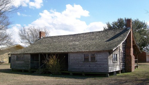 A rustic wooden house with a sloped roof, two chimneys, and a grassy yard under a partly cloudy sky.