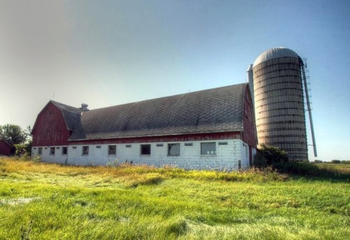 A red barn with a silo stands in a grassy field under a clear blue sky.
