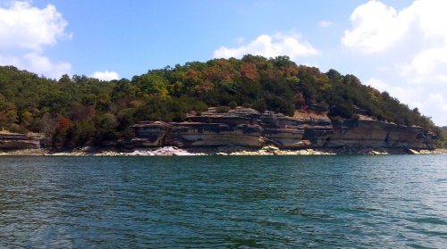 A scenic view of a rocky shoreline with lush greenery and colorful trees under a blue sky with fluffy clouds.