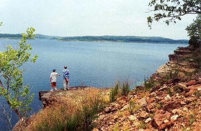 Two people stand on a rocky ledge overlooking a calm lake surrounded by hills and trees under a clear sky.