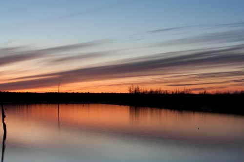 A serene sunset over a calm lake, with colorful clouds reflecting on the water and silhouettes of trees in the background.