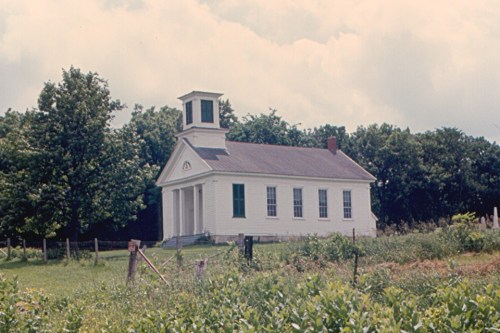A white, historic schoolhouse with a bell tower, surrounded by green fields and trees under a cloudy sky.